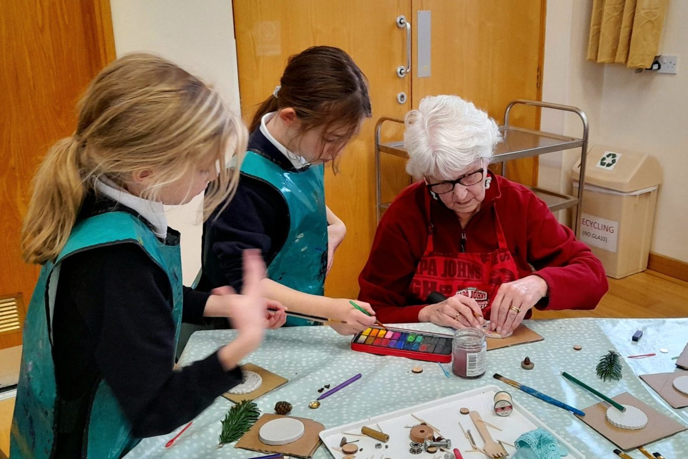 Two children craft with an elderly woman, surrounded by materials on a table.
